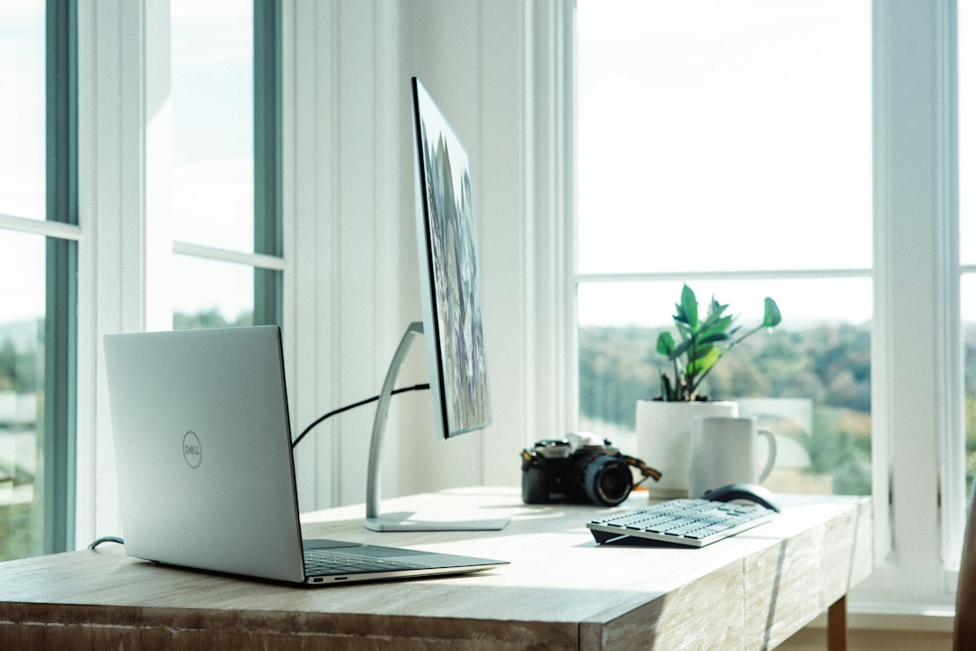 A laptop, monitor, camera, and plant on a wooden desk in a sunlit office