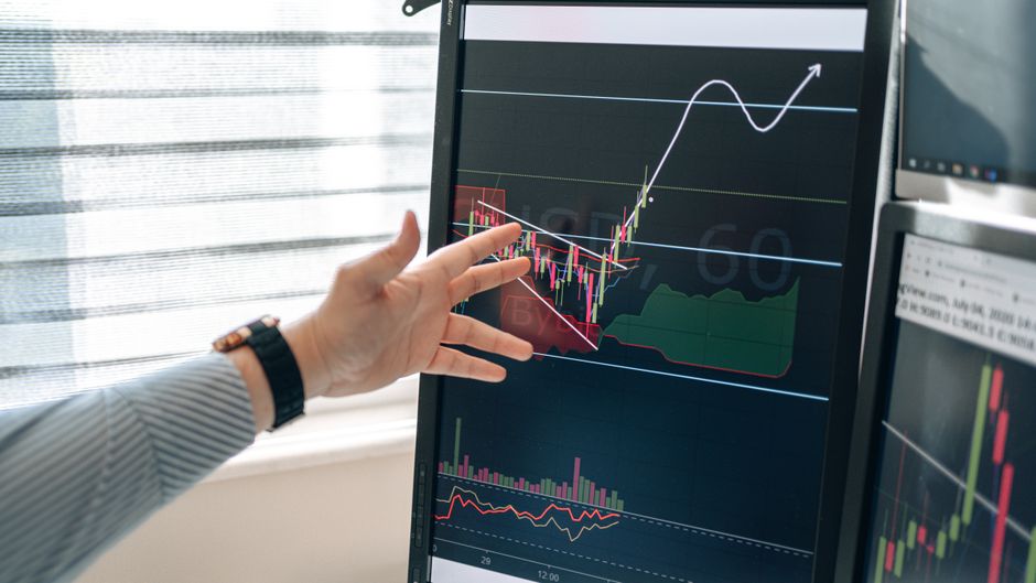 Close-up of a hand pointing at stock market graphs on a monitor in a workspace