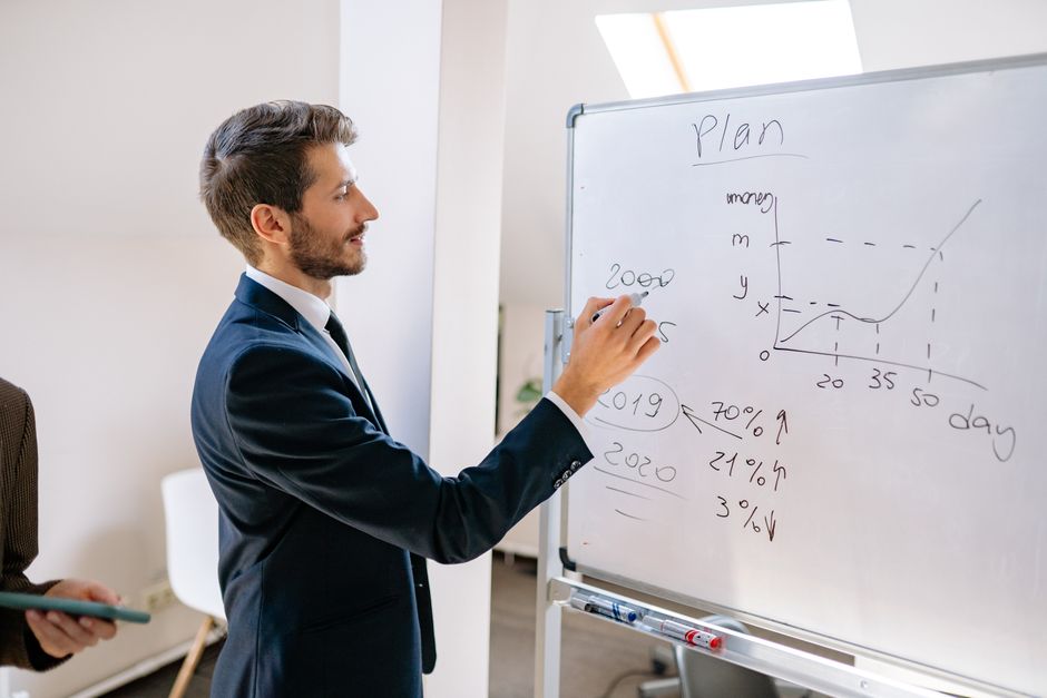 A businessman in a suit writes financial data on a whiteboard during an office planning session