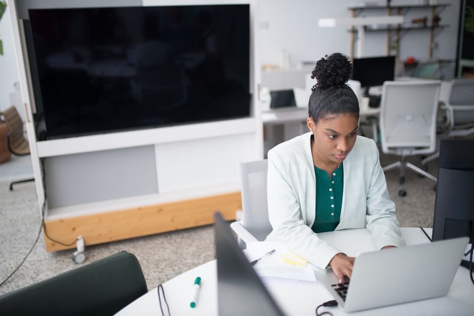 Focused woman using a laptop in a modern office setting, wearing a white blazer, working diligently