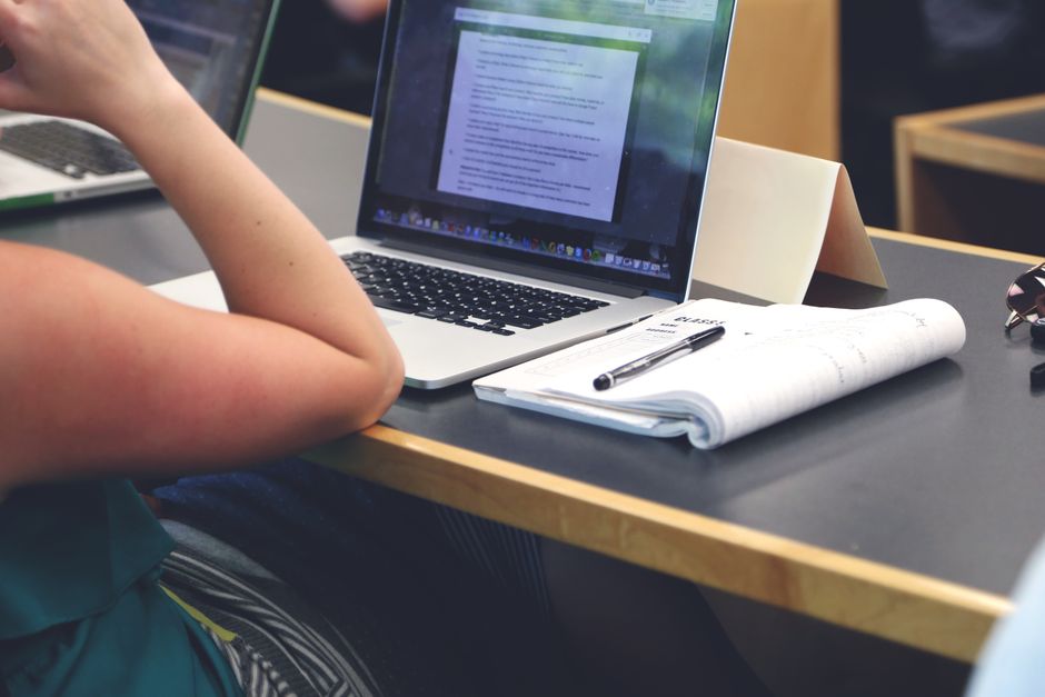A student in a classroom setting using a laptop and taking notes in a notebook