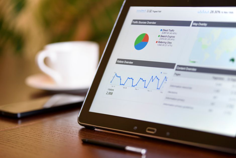 Close-up of a tablet displaying analytics charts on a wooden office desk, alongside a smartphone and coffee cup