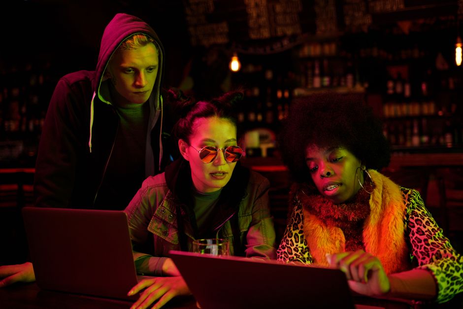 Three friends collaborating over laptops in a dimly lit pub setting, showcasing diversity and teamwork
