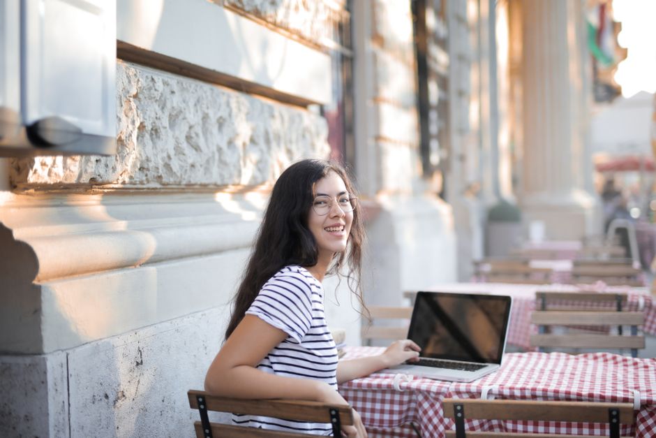 Happy woman with long hair and glasses sitting outdoors, using a laptop and smiling