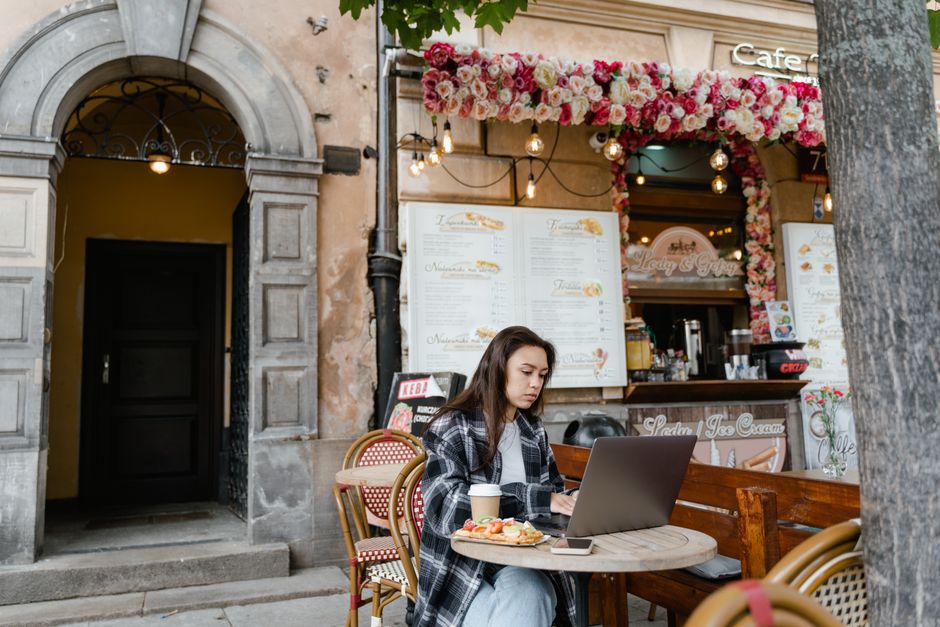 A woman at an outdoor cafe working on a laptop with coffee and snacks