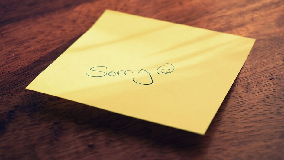 A yellow sticky note with a handwritten apology message and smiley face on a wooden surface
