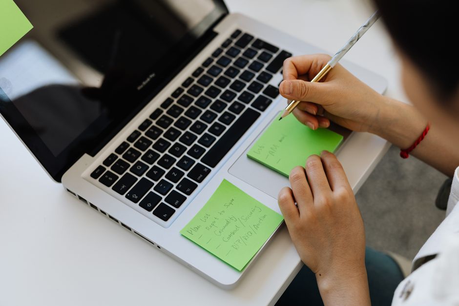 An individual writes notes on green sticky notes placed on a laptop in a work setting